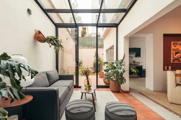 Modern living room with gray sofa, glass skylight ceiling, potted plants, and open courtyard view beyond black-framed doors